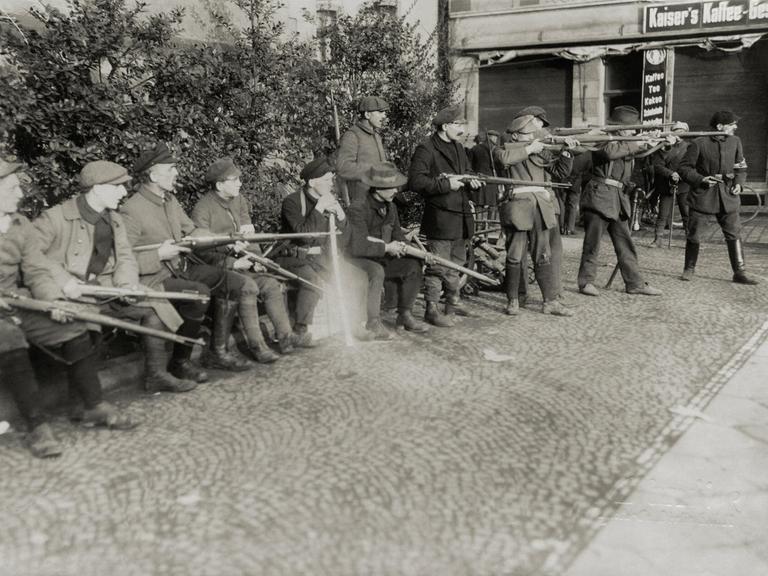 Schwarz-weißes Archivfoto: In einer Stadt im Ruhrgebiet 1920 stehen mehrere Männer nebeneinander. Sie haben alle Gewehre bei sich, einige davon schießen. Schwarz-weißes Archivfoto: In einer Stadt im Ruhrgebiet 1920 stehen mehrere Männer nebeneinander. Sie haben alle Gewehre bei sich, einige davon schießen.