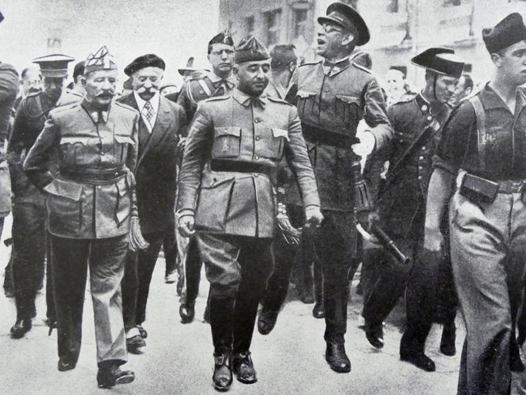 Schwarz-Weiß-Fotografie aus dem August 1936: General Francisco Franco schreitet in Burgos flankiert von den Generälen Emilio Mola, Miguel Cabanellas und José Cavalcanti durch eine Straße. Schwarz-Weiß-Fotografie aus dem August 1936: General Francisco Franco schreitet in Burgos flankiert von den Generälen Emilio Mola, Miguel Cabanellas und José Cavalcanti durch eine Straße.