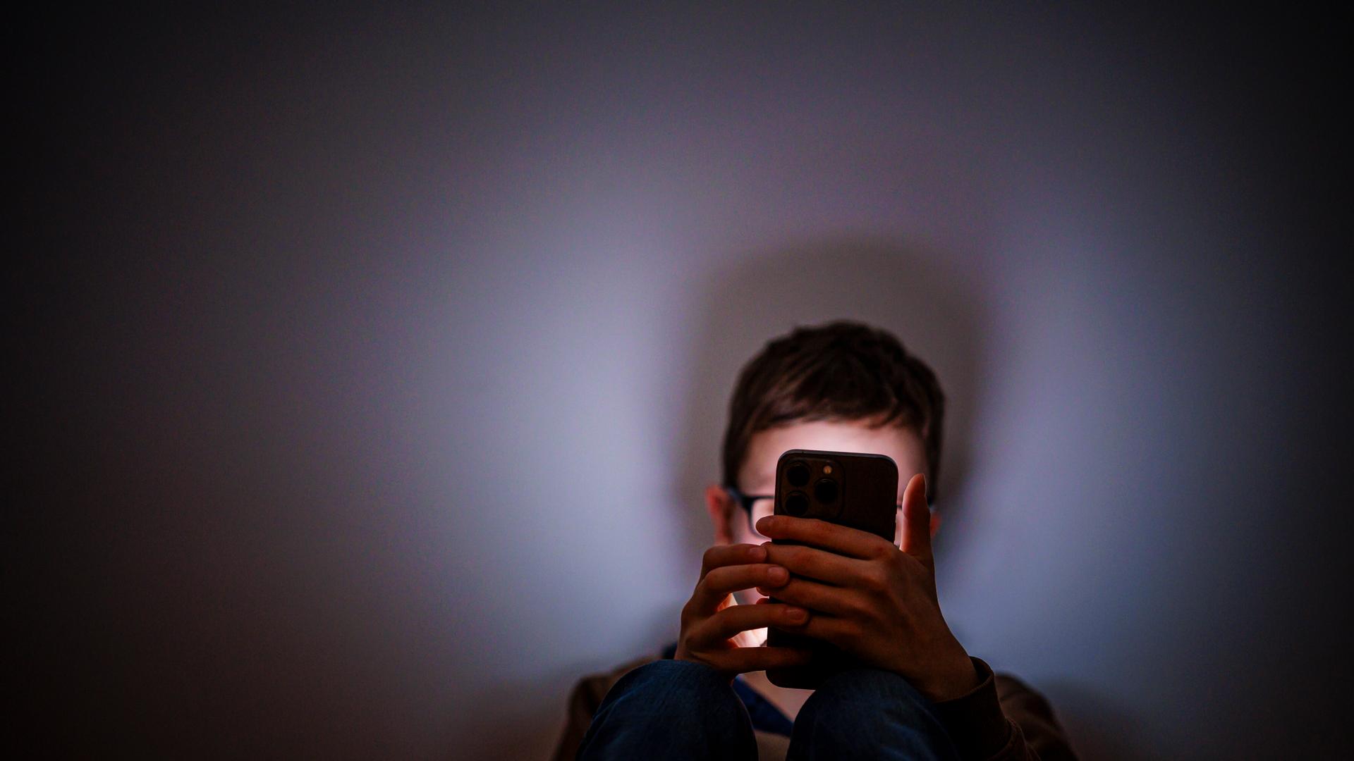 Ein Junge sitzt in einem dunklen Raum und wird von einem Smartphone beleuchtet. Ein Junge sitzt in einem dunklen Raum und wird von einem Smartphone beleuchtet.