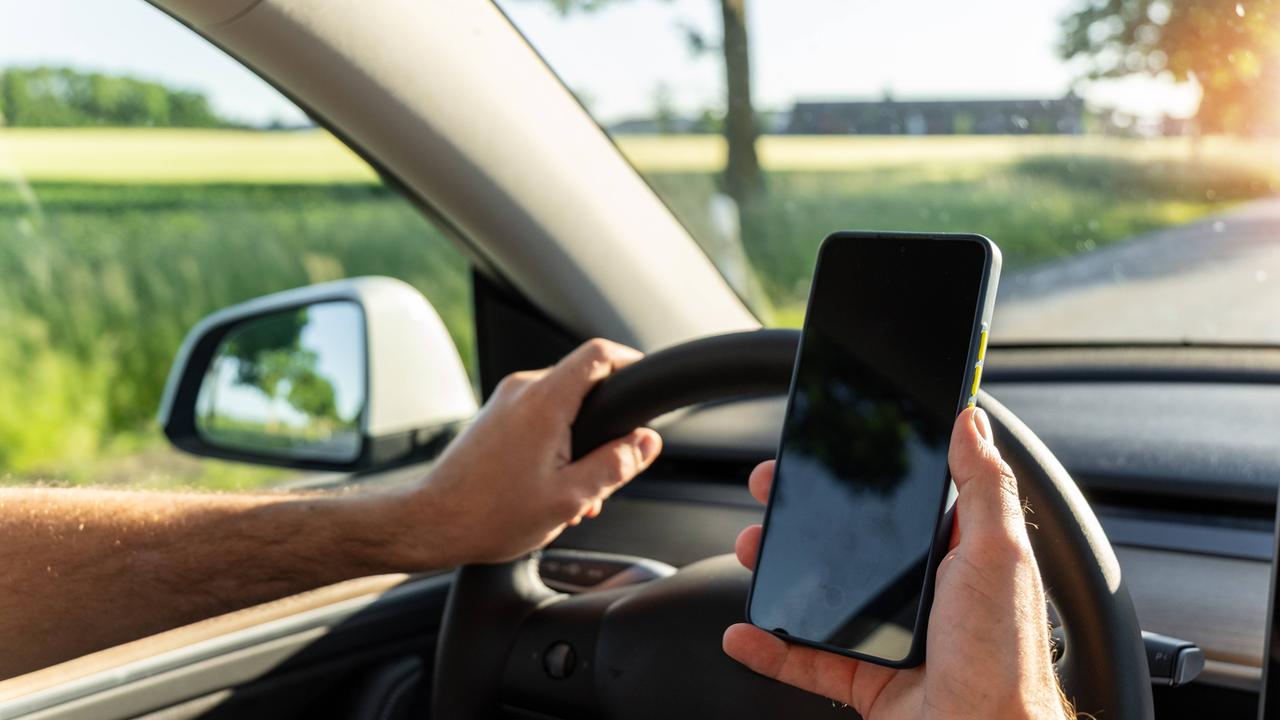 Ein Mann sitzt am Steuer eines fahrenden Autos und hält ein Smartphone in der Hand.