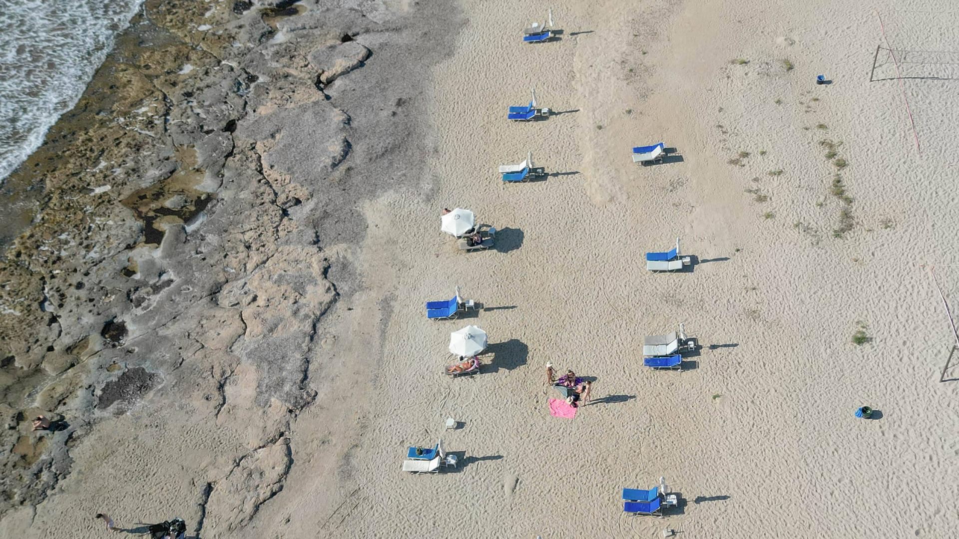 Blick aus der Vogelperspektive: Auf Zypern stehen am Strand Liegestühle mit Sonnenschirmen, es sind wenige Menschen am Strand. Es wirkt sehr heiß.