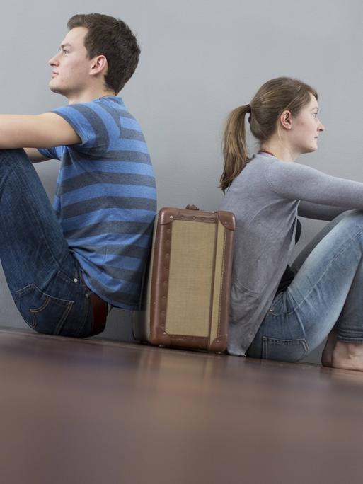 Mann und Frau sitzen mit dem Rücken zueinander auf dem Boden, zwischen ihnen steht ein Koffer