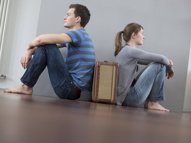 Mann und Frau sitzen mit dem Rücken zueinander auf dem Boden, zwischen ihnen steht ein Koffer
