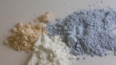 Auf einem Tisch liegen drei Pulverhaufen in braun, weiß und blau. Es sind die Seltenen Erden Ceroxid, Yttriumoxid und Neodymoxid.