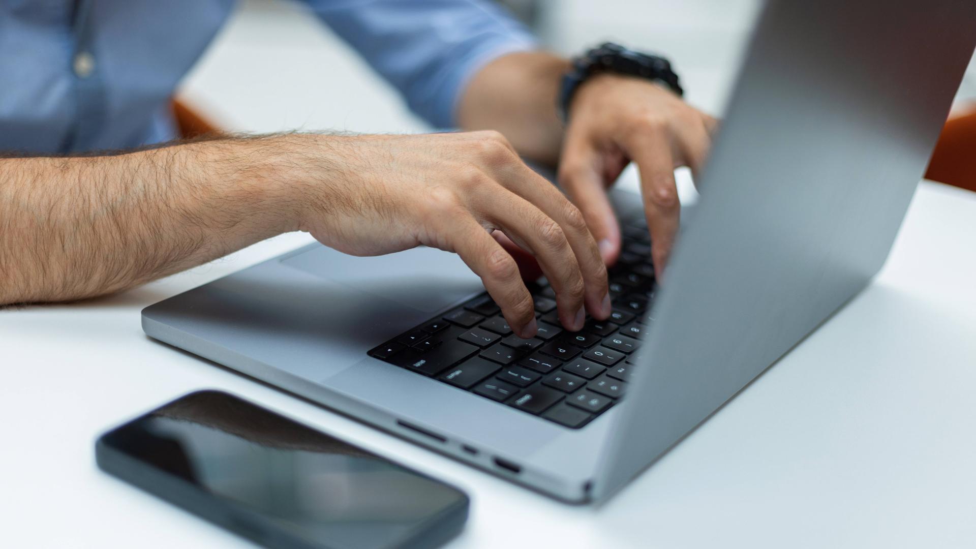 Nahaufnahme der Hände eines Mannes, die auf der Tastatur eines Laptops an einem weißen Schreibtisch tippen.