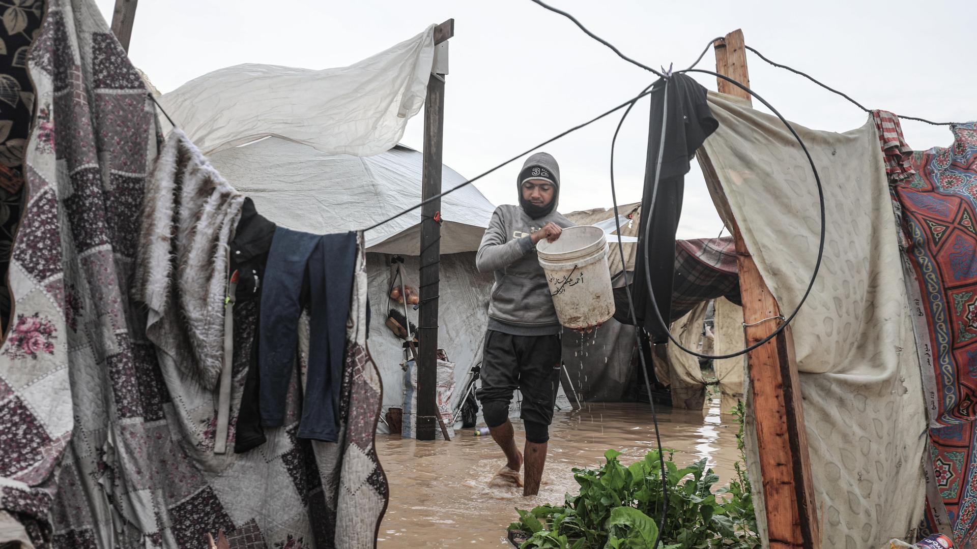 Mann, der einen Eimer hält, geht auf einem überfluteten Platz an Zelten vorbei (12.12.2025, Deir Al-Balah in Gaza)