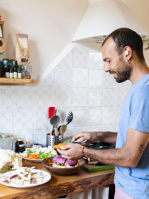 Ein Mann steht lächelnd in einer Küche und bereitet dabei Gemüse zu. Er hat einen kabellosen Kopfhörer im Ohr.