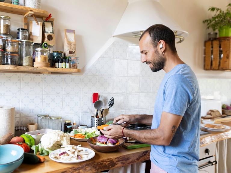 Ein Mann steht lächelnd in einer Küche und bereitet dabei Gemüse zu. Er hat einen kabellosen Kopfhörer im Ohr.