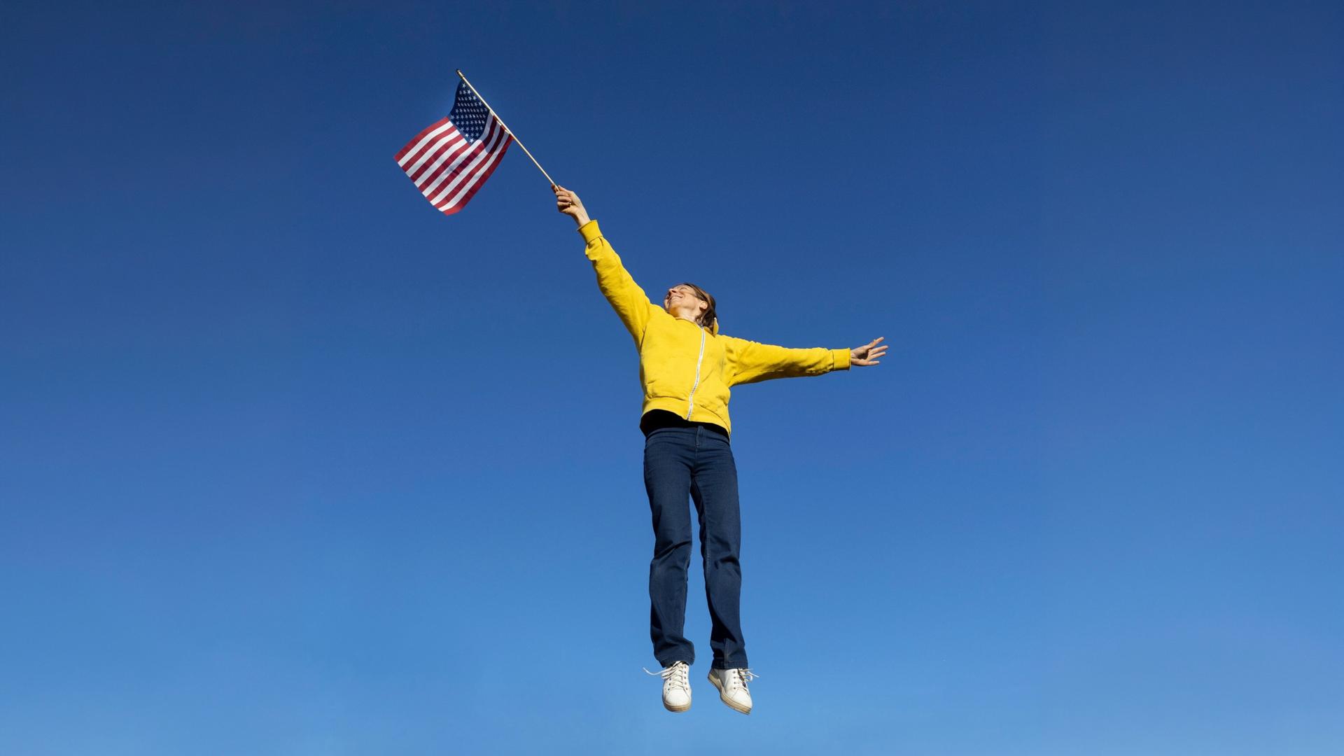 Eine Frau trägt ein gelbes Sweatshirt, eine dunkle Jeans und hält die amerikanische Fahne hoch mit einem ausgestreckten Arm und springt dabei in die Luft vor einem blauen Himmel.