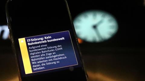 Eine Warnmeldung auf einem Smartphone über eine bundesweite IT-Störung im Bahnverkehr. Im Hintergrund eine Bahnhofsuhr, die ein Uhr fünfundzwanzig in der Nacht anzeigt.