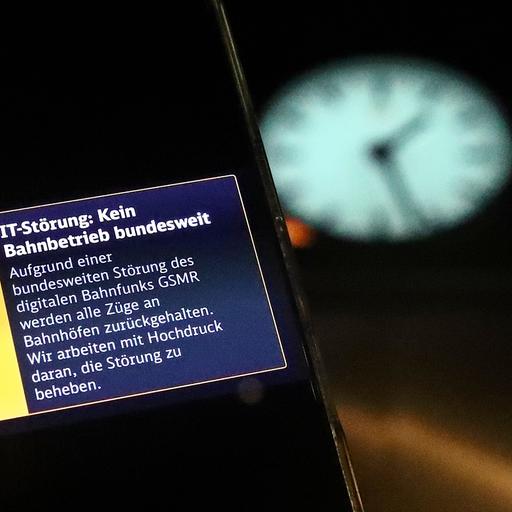 Eine Warnmeldung auf einem Smartphone über eine bundesweite IT-Störung im Bahnverkehr. Im Hintergrund eine Bahnhofsuhr, die ein Uhr fünfundzwanzig in der Nacht anzeigt.