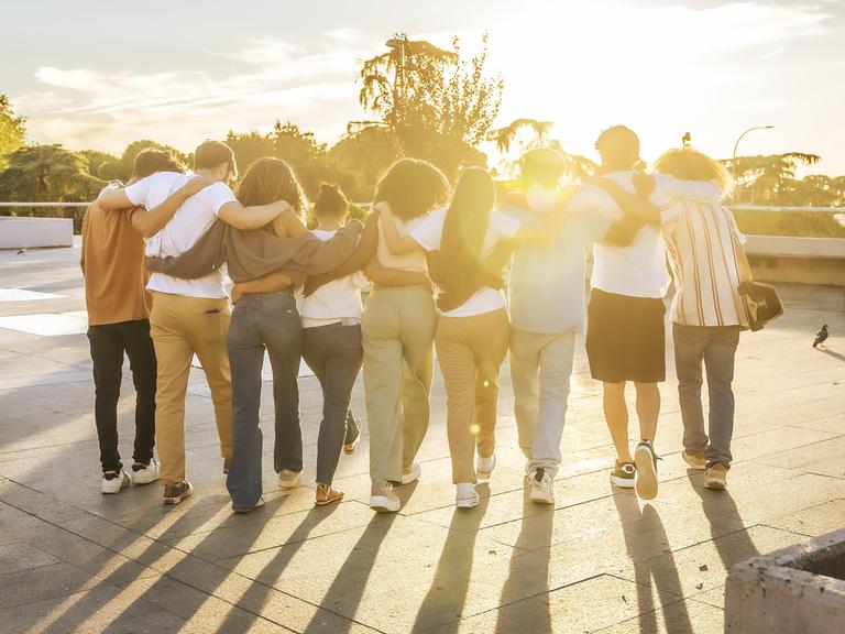 Rückansicht von Freunden, die sich umarmen und bei Sonnenuntergang gemeinsam durch die Stadt laufen