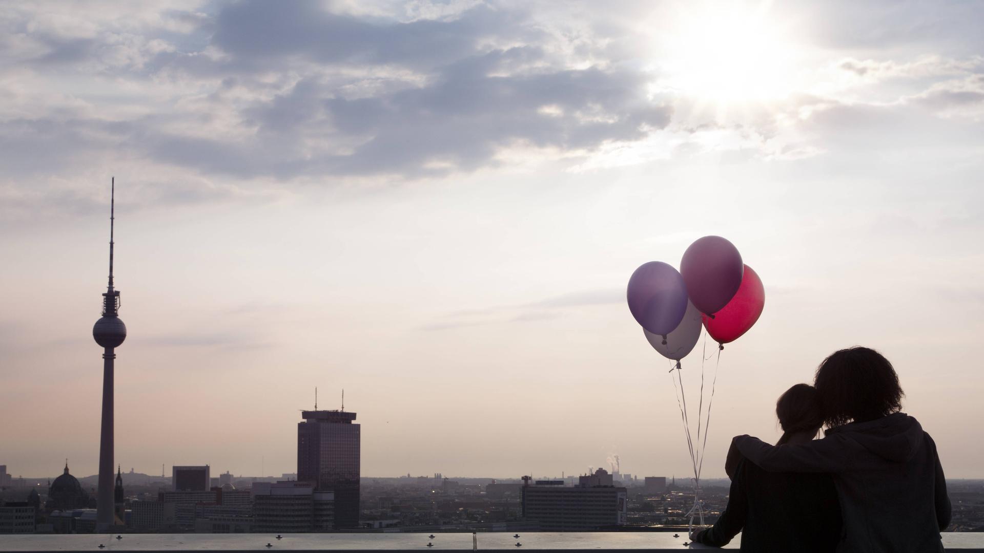 Zwei Personen stehen auf einer Dachterrasse und blicken auf Berlin, die Sonne scheint. Sie halten vier bunte Luftballons in der Hand.