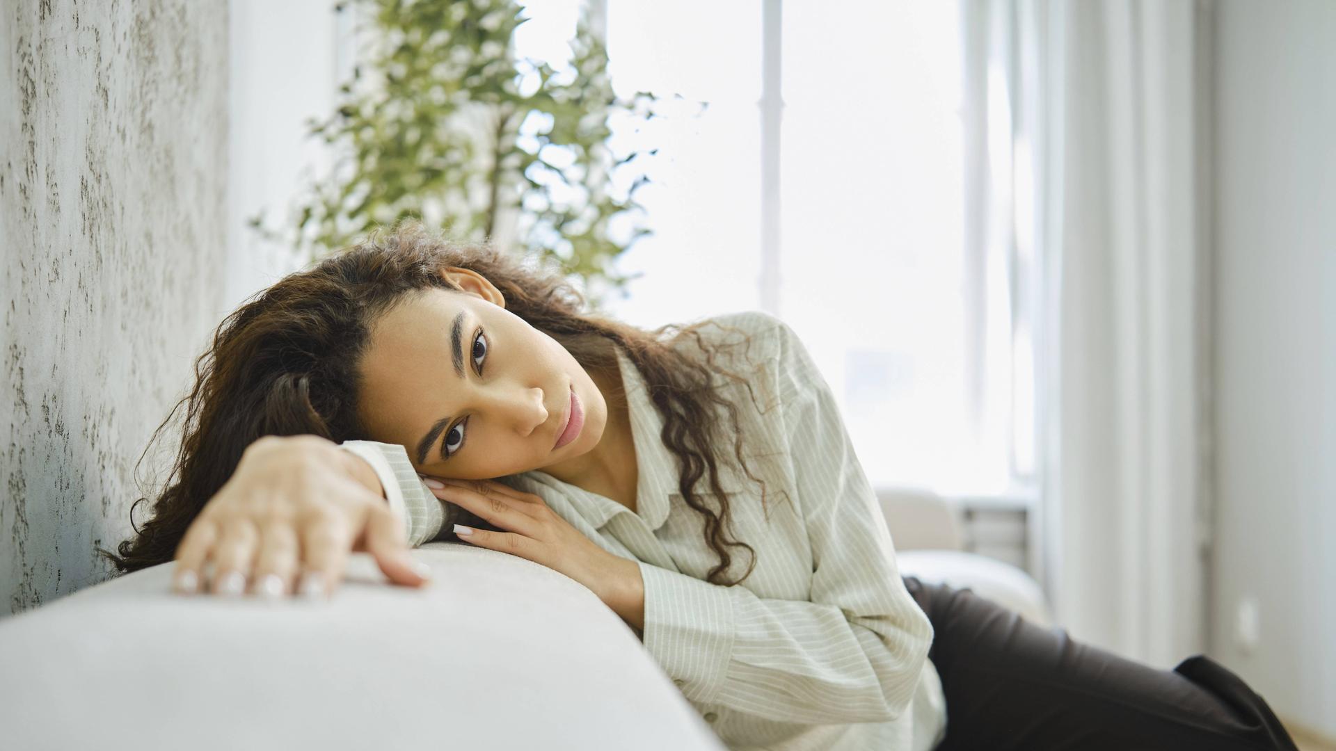 Eine Frau mit lockigem Haar lehnt auf einer Couch in einem hellen Wohnzimmer. Sie wirkt nachdenklich. Eine Frau mit lockigem Haar lehnt auf einer Couch in einem hellen Wohnzimmer. Sie wirkt nachdenklich.