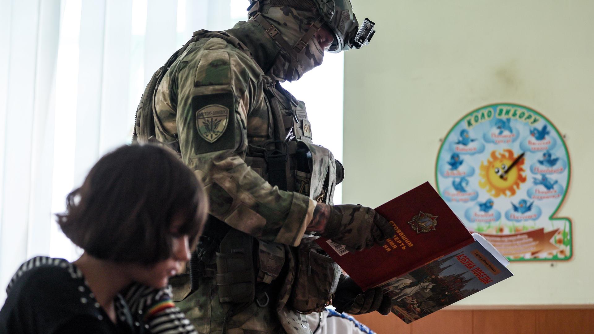 Ein Militär geht mit einem Buch an einem im Klassenraum sitzenden Schulkind vorbei (Kurakhovo, ukrainische und von Russland besetzte Region Donezk, 23.09.2025)
