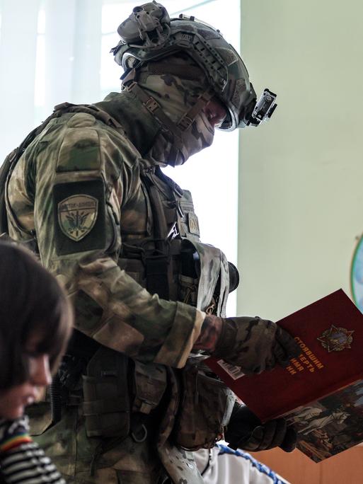 Ein Militär geht mit einem Buch an einem im Klassenraum sitzenden Schulkind vorbei (Kurakhovo, ukrainische und von Russland besetzte Region Donezk, 23.09.2025)