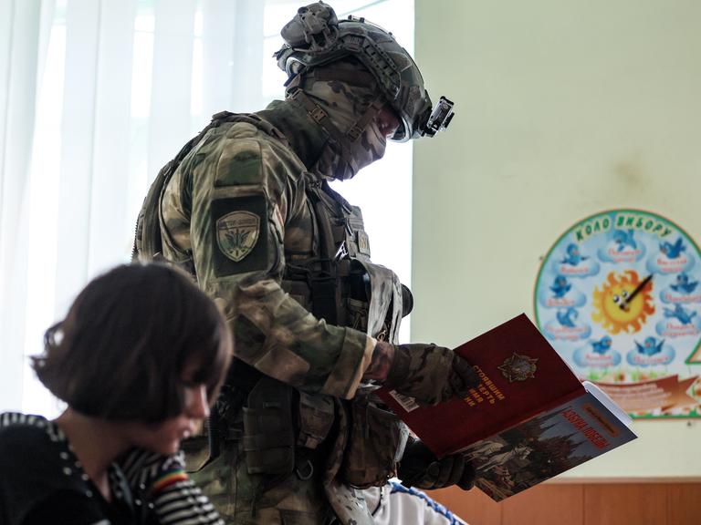Ein Militär geht mit einem Buch an einem im Klassenraum sitzenden Schulkind vorbei (Kurakhovo, ukrainische und von Russland besetzte Region Donezk, 23.09.2025)