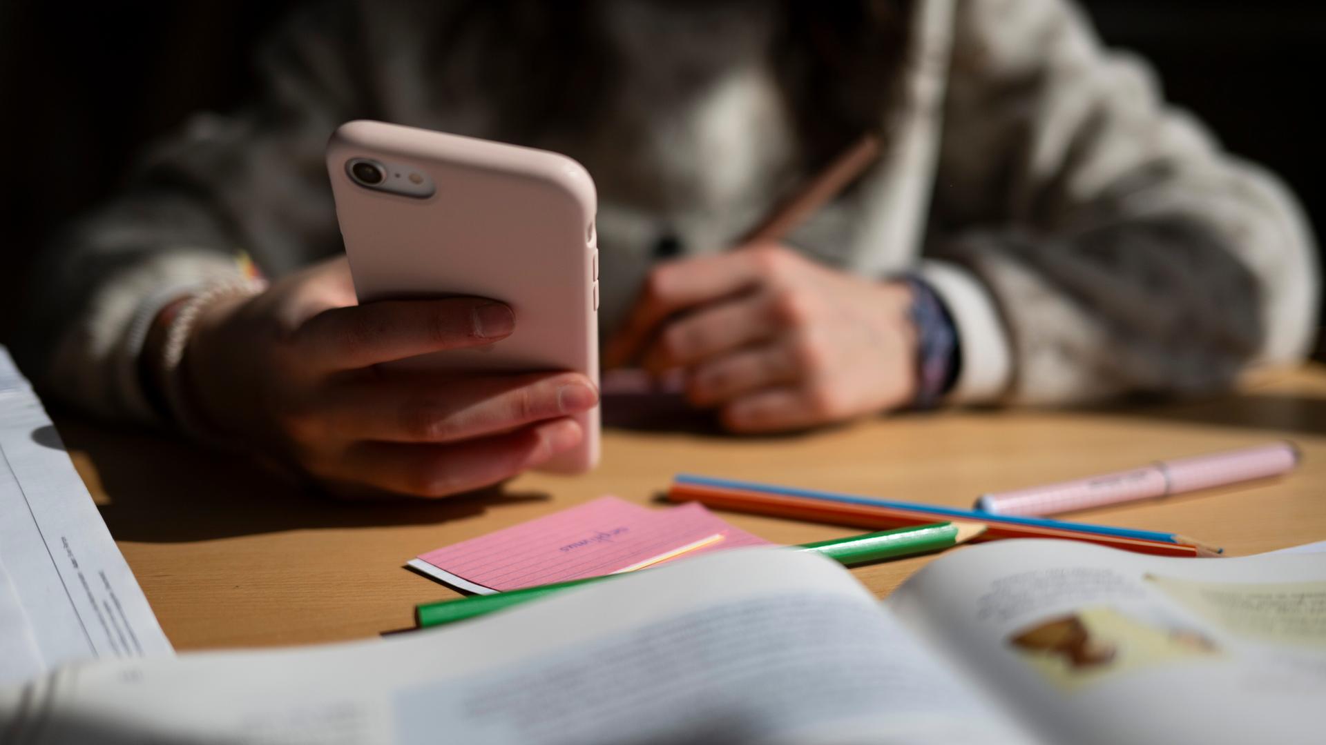Ein Schulkind sitzt am Schreibtisch, bearbeitet Aufgaben mit einem Stift und hält dabei ein Smartphone in der Hand. Ein Schulkind sitzt am Schreibtisch, bearbeitet Aufgaben mit einem Stift und hält dabei ein Smartphone in der Hand.