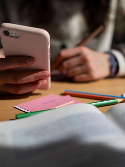 Ein Schulkind sitzt am Schreibtisch, bearbeitet Aufgaben mit einem Stift und hält dabei ein Smartphone in der Hand. Ein Schulkind sitzt am Schreibtisch, bearbeitet Aufgaben mit einem Stift und hält dabei ein Smartphone in der Hand.