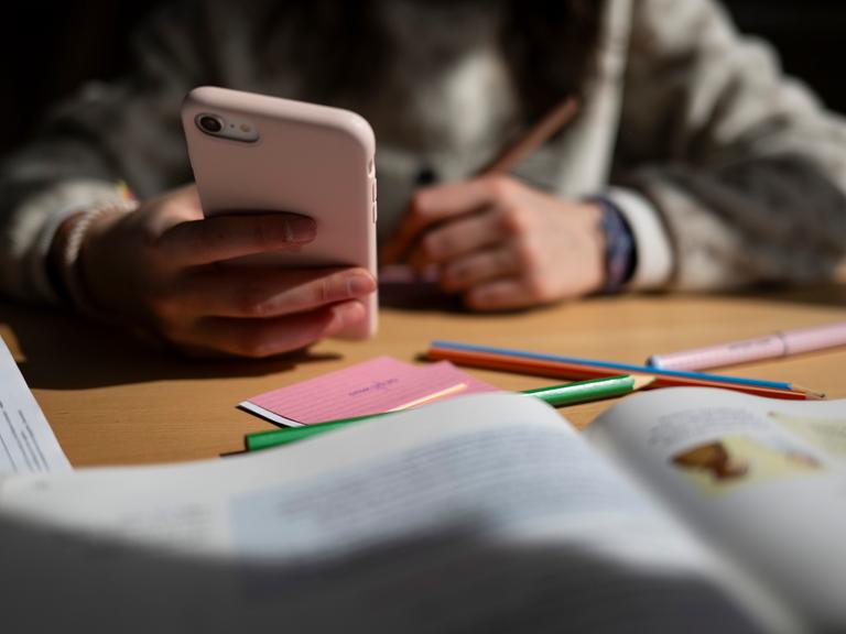 Ein Schulkind sitzt am Schreibtisch, bearbeitet Aufgaben mit einem Stift und hält dabei ein Smartphone in der Hand. Ein Schulkind sitzt am Schreibtisch, bearbeitet Aufgaben mit einem Stift und hält dabei ein Smartphone in der Hand.