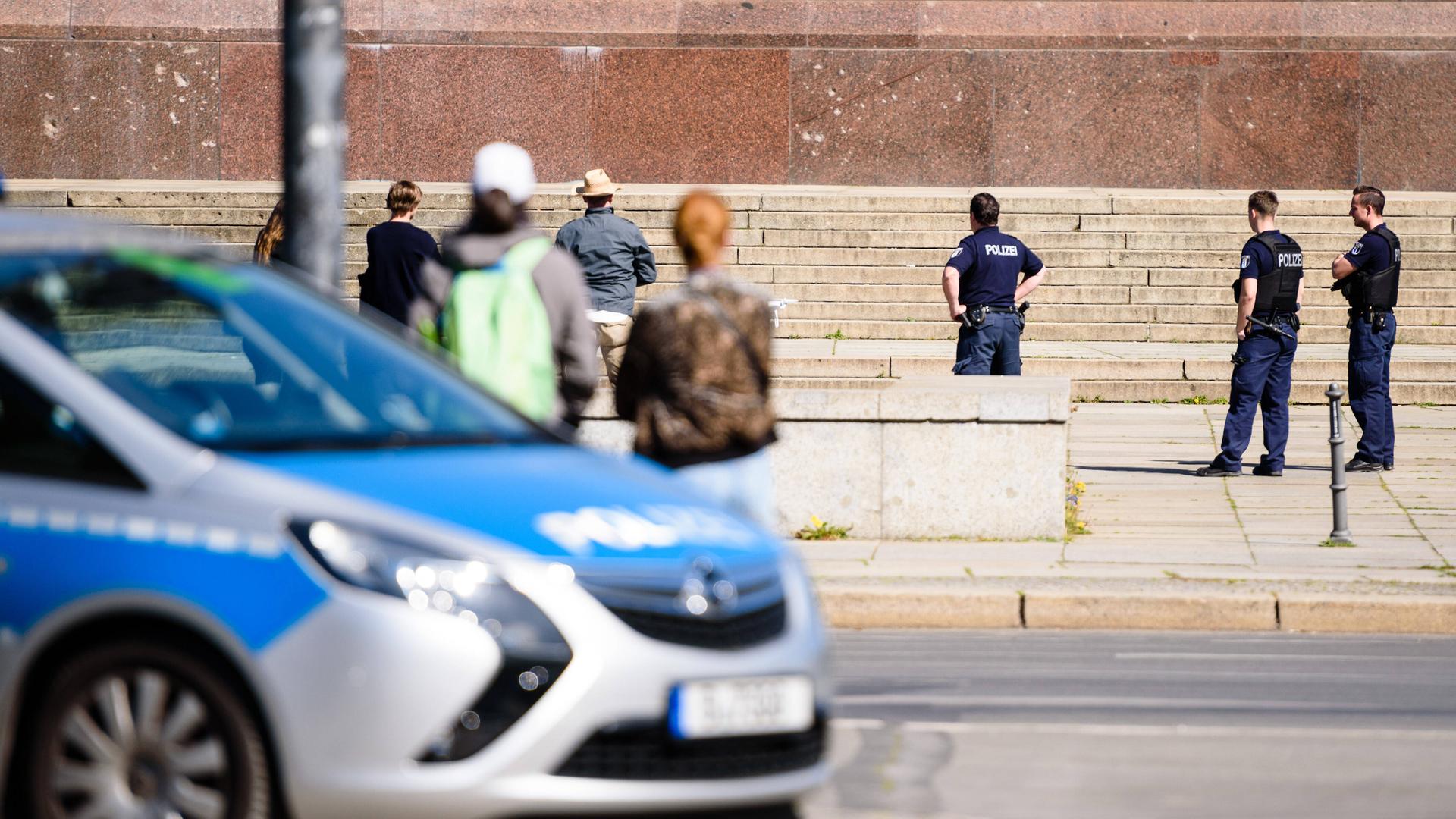 An einer Straßenecke parkt ein Polizeiauto. Im Hintergrund stehen mehrere Polizisten auf einem Platz und schauen auf eine Drone. An einer Straßenecke parkt ein Polizeiauto. Im Hintergrund stehen mehrere Polizisten auf einem Platz und schauen auf eine Drone.