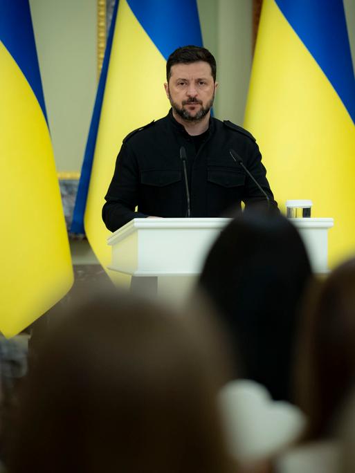Der ukrainische Präsident Volodymyr Selenskyj steht am Mikrofon, hinter stehen ukrainische Flaggen in einer langen Reihe
