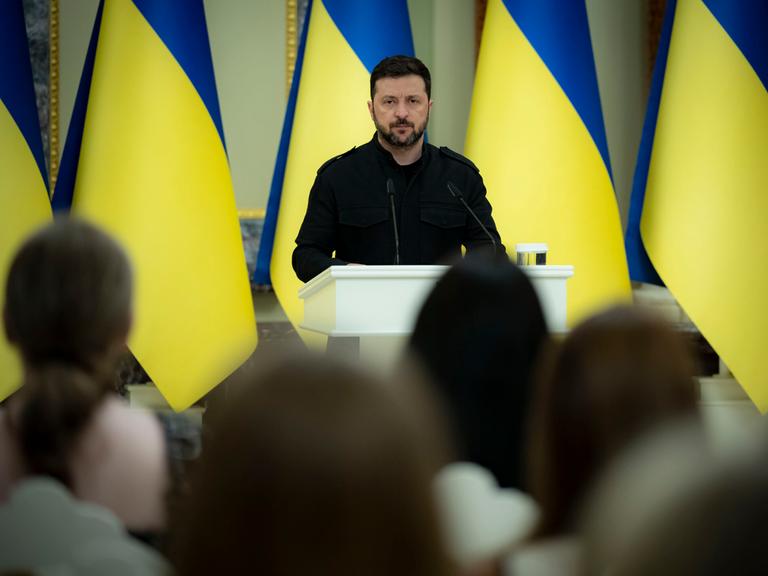 Der ukrainische Präsident Volodymyr Selenskyj steht am Mikrofon, hinter stehen ukrainische Flaggen in einer langen Reihe