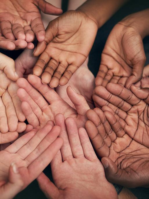 Viele Hände mit verschiedenen Hautfarben liegen übereinander, es wirkt nach Gemeinschaft.