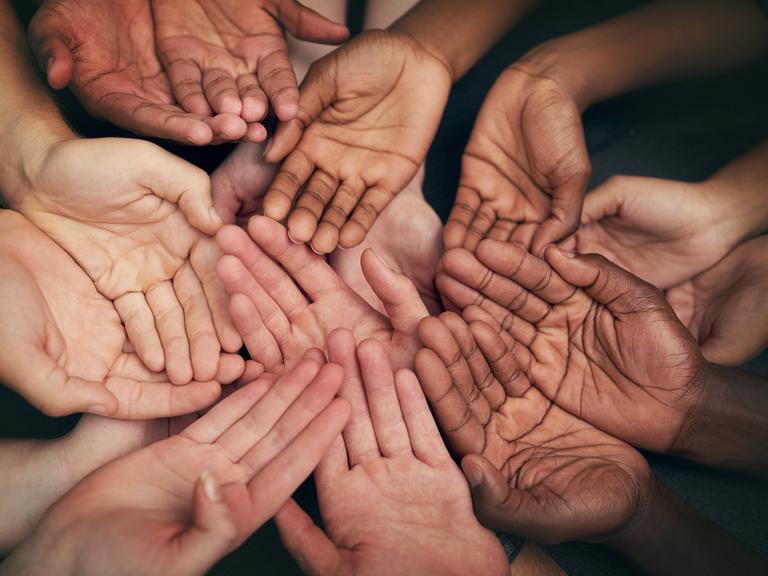 Viele Hände mit verschiedenen Hautfarben liegen übereinander, es wirkt nach Gemeinschaft.