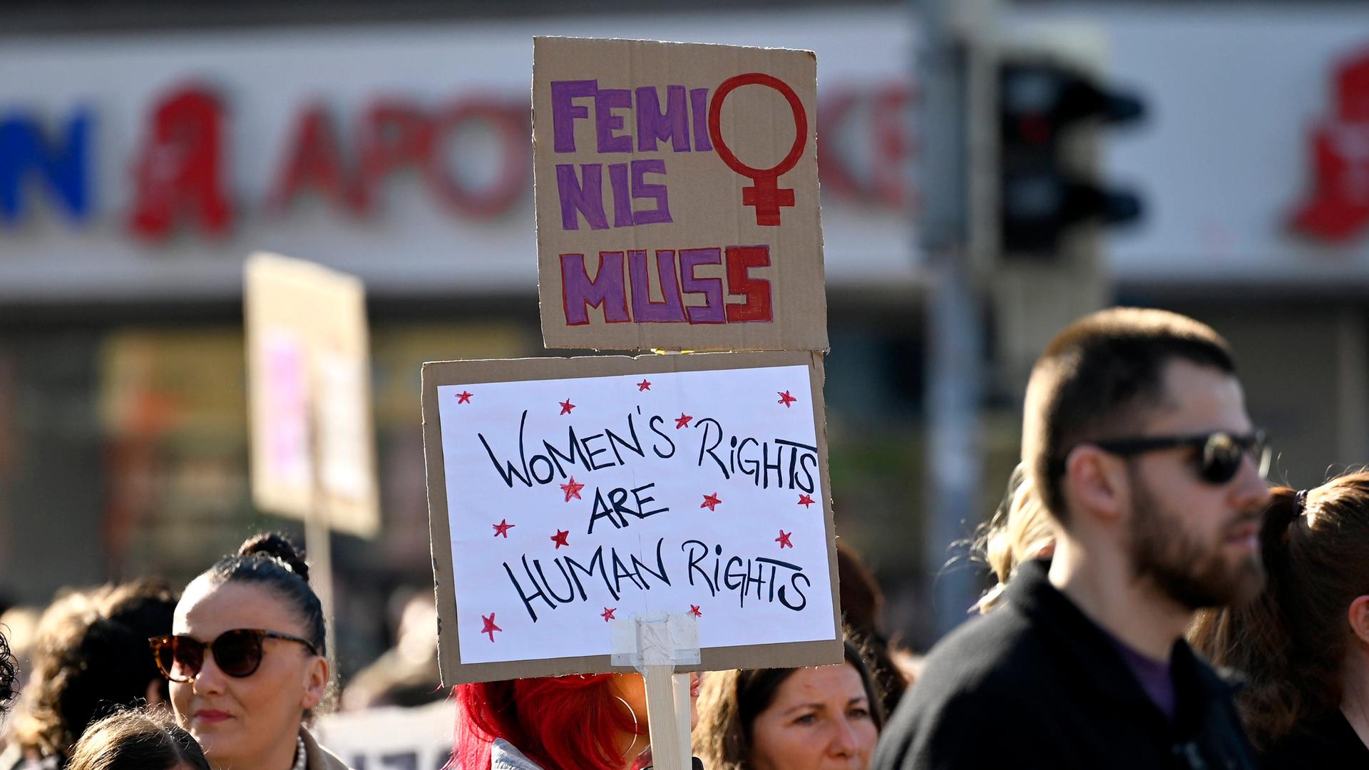 Auf einer Demo zum Weltfrauentag hält eine Teilnehmerin zwei Schilder in die Höhe, auf dem unteren steht "Women’s rights are human rights", auf dem oberen Feminismus mit zwei s (Frankfurt, 08.03.2025) Auf einer Demo zum Weltfrauentag hält eine Teilnehmerin zwei Schilder in die Höhe, auf dem unteren steht "Women’s rights are human rights", auf dem oberen Feminismus mit zwei s (Frankfurt, 08.03.2025)