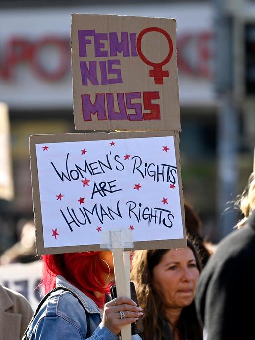 Auf einer Demo zum Weltfrauentag hält eine Teilnehmerin zwei Schilder in die Höhe, auf dem unteren steht "Women’s rights are human rights", auf dem oberen Feminismus mit zwei s (Frankfurt, 08.03.2025)