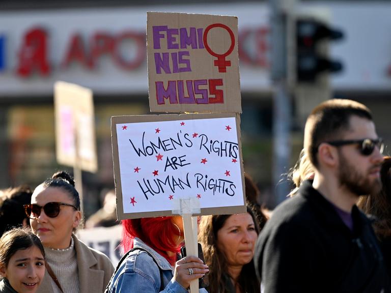 Auf einer Demo zum Weltfrauentag hält eine Teilnehmerin zwei Schilder in die Höhe, auf dem unteren steht "Women’s rights are human rights", auf dem oberen Feminismus mit zwei s (Frankfurt, 08.03.2025)