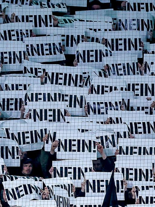 Fußballfans halten Schilder als Protest gegen die Pläne des Bundesinnenministers mit der Aufschrift „NEIN“ vor ihre Gesichter (Borussia-Park Mönchengladbach, 28.11.2025)