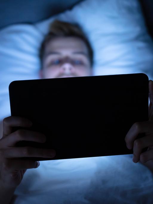 Foto eines Mannes, der auf dem Rücken im Bett liegt und auf einem Tablet einen Film schaut. Foto eines Mannes, der auf dem Rücken im Bett liegt und auf einem Tablet einen Film schaut.