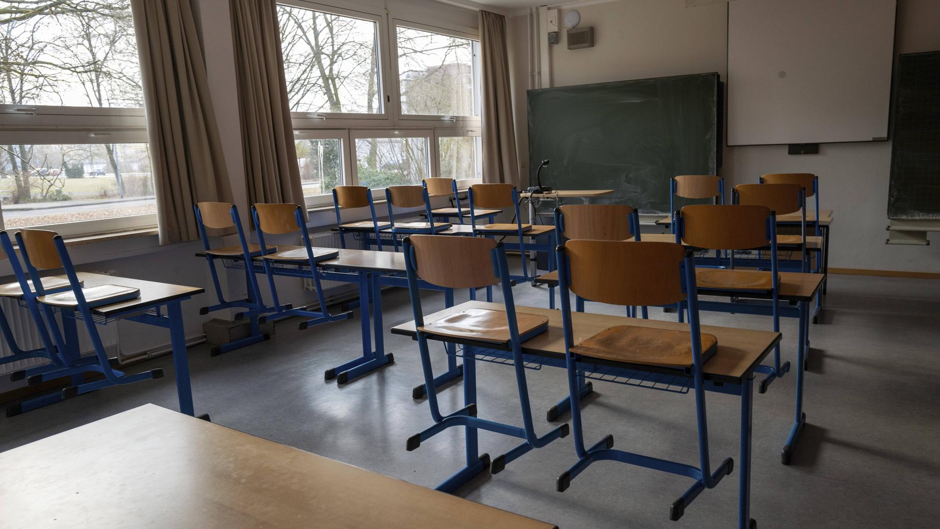 Ein leerer, tageslichtdurchfluteter Klassenraum mit hölzernen Schultischen und Stühlen mit blauen Metallbeinen. Im Hintergrund befinden sich eine Kreidetafel und eine weiße Projektionsfläche an der Wand.