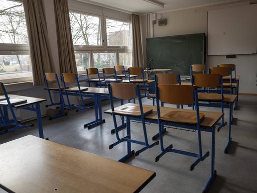 Ein leerer, tageslichtdurchfluteter Klassenraum mit hölzernen Schultischen und Stühlen mit blauen Metallbeinen. Im Hintergrund befinden sich eine Kreidetafel und eine weiße Projektionsfläche an der Wand. Ein leerer, tageslichtdurchfluteter Klassenraum mit hölzernen Schultischen und Stühlen mit blauen Metallbeinen. Im Hintergrund befinden sich eine Kreidetafel und eine weiße Projektionsfläche an der Wand.