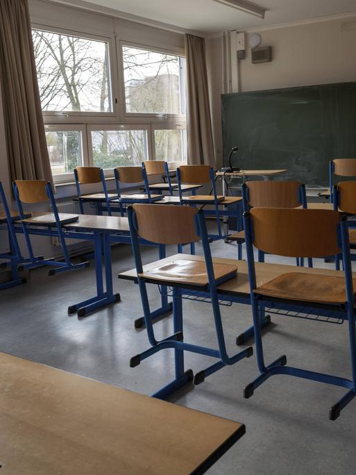 Ein leerer, tageslichtdurchfluteter Klassenraum mit hölzernen Schultischen und Stühlen mit blauen Metallbeinen. Im Hintergrund befinden sich eine Kreidetafel und eine weiße Projektionsfläche an der Wand. Ein leerer, tageslichtdurchfluteter Klassenraum mit hölzernen Schultischen und Stühlen mit blauen Metallbeinen. Im Hintergrund befinden sich eine Kreidetafel und eine weiße Projektionsfläche an der Wand.