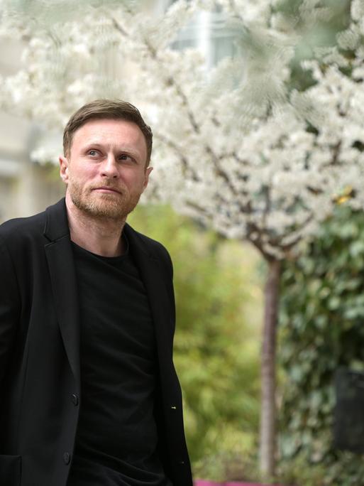 Der deutsche Autor Michael Nast steht draußen in einem Garten, er trägt ein schwarzes Jackett und Oberteil. Im Hintergrund blüht ein weißer Kirschbaum