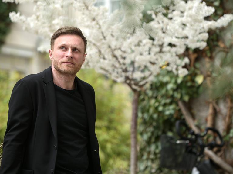 Der deutsche Autor Michael Nast steht draußen in einem Garten, er trägt ein schwarzes Jackett und Oberteil. Im Hintergrund blüht ein weißer Kirschbaum