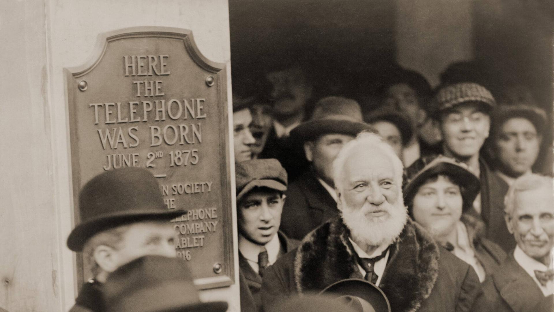 Schwarz-weiß Archifoto: Der Erfinder Graham Bell steht inmitten einer Menschengruppe. An der Wand neben ihm wurde ein Schild angebracht, das ihn als Erfinder des Telefons ehrt.