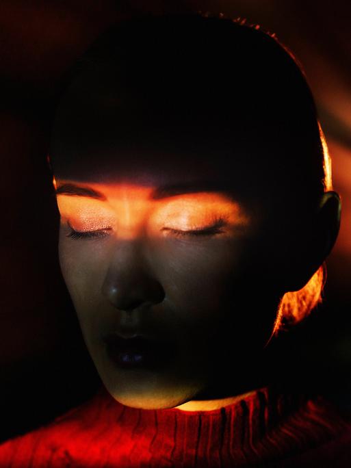 Eine Frau ist alleine in einem abgedunkelten Raum. Hinter ihr leuchtet ein Licht. Sie hat ihre Augen geschlossen. Auf ihre Augen leuchtet ein Lichtstrahl (Symbolbild) Eine Frau ist alleine in einem abgedunkelten Raum. Hinter ihr leuchtet ein Licht. Sie hat ihre Augen geschlossen. Auf ihre Augen leuchtet ein Lichtstrahl (Symbolbild)