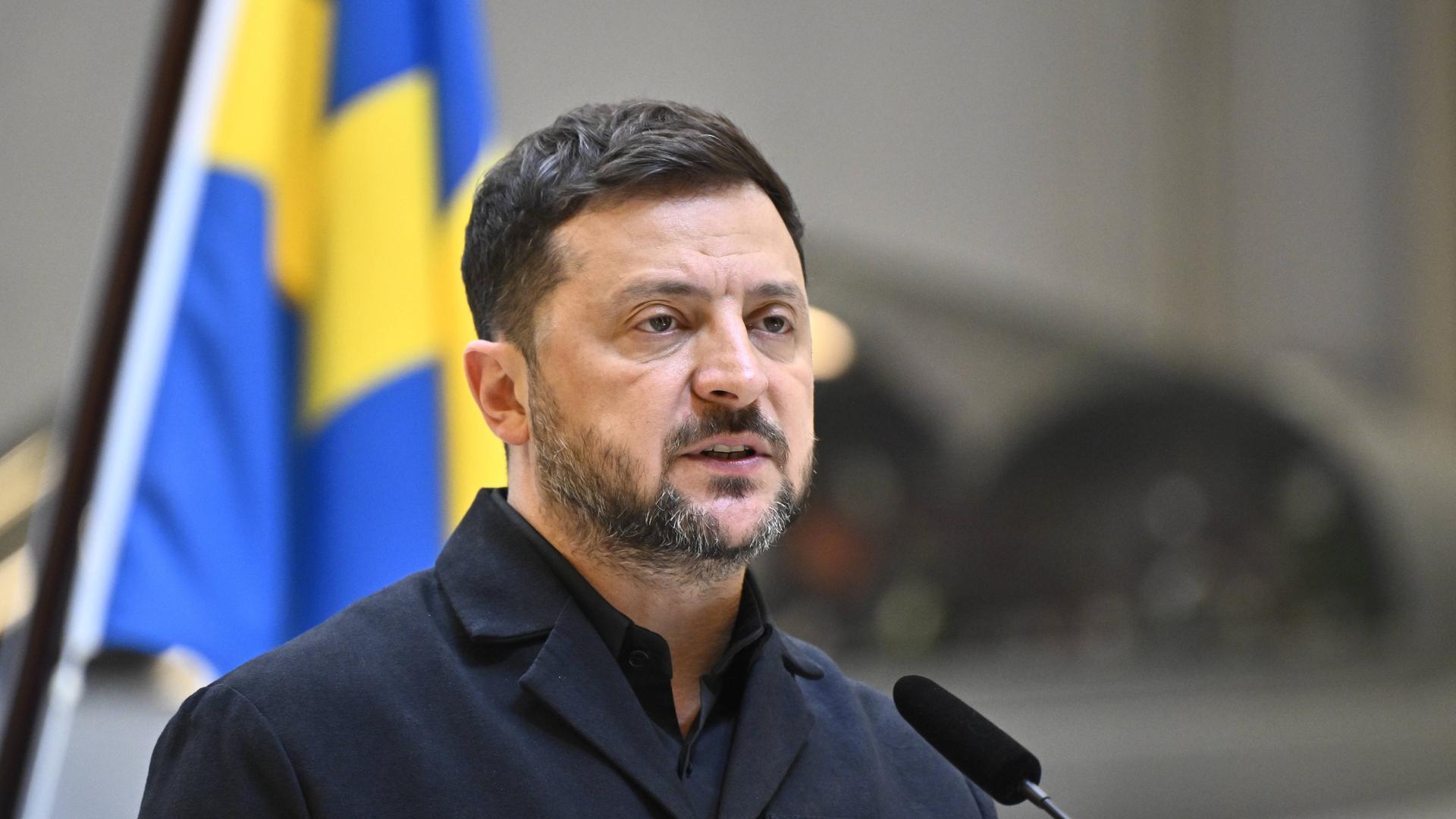 Der ukrainische Präsident Wolodymyr Selenskyj steht auf einer Pressekonferenz vor der Flagge seines Landes und spricht in ein Mikrofon.