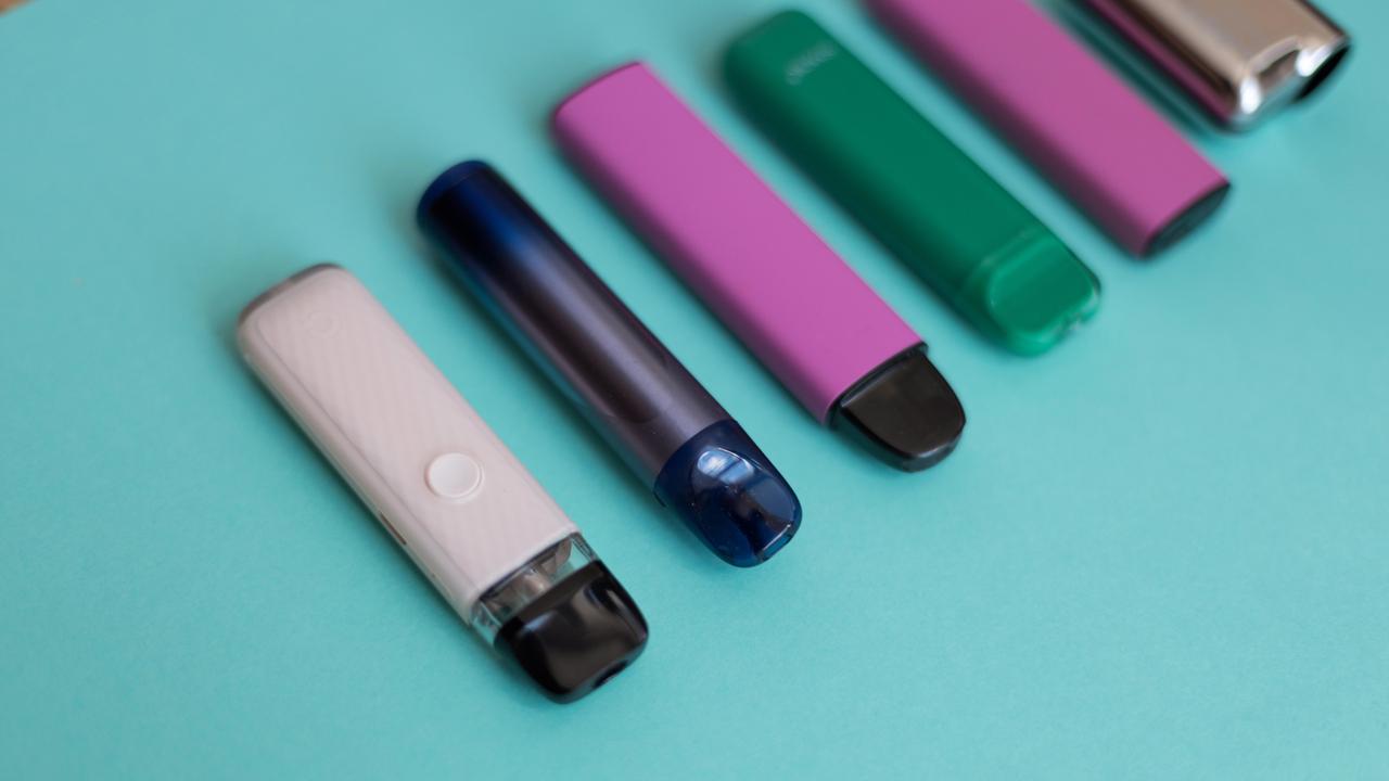 E-Zigaretten in den Farben Grün, Rosa, Dunkelblau und Beige liegen nebeneinander auf einem grünen Hintergrund.