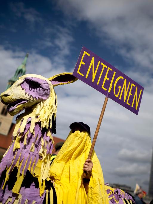 Demonstrant der Initiative Volksentscheid Deutsche Wohnen und Co ist als lila-gelbes Einhorn oder Trojanisches Pferd verkleidet und hält ein Schild mit dem Wort "Enteignung" in die Höhe (05.09.2026, Berlin)