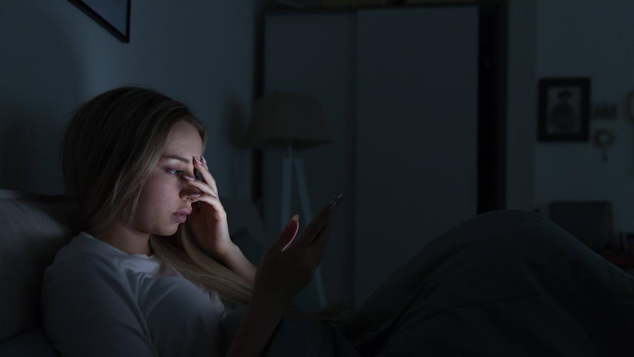 Eine junge Frau liegt in einem dunklen Zimmer in einem Bett. Das Licht ihres Handydisplays erhellt ihr Gesicht. Sie hält sich eine Hand vor das Gesicht.