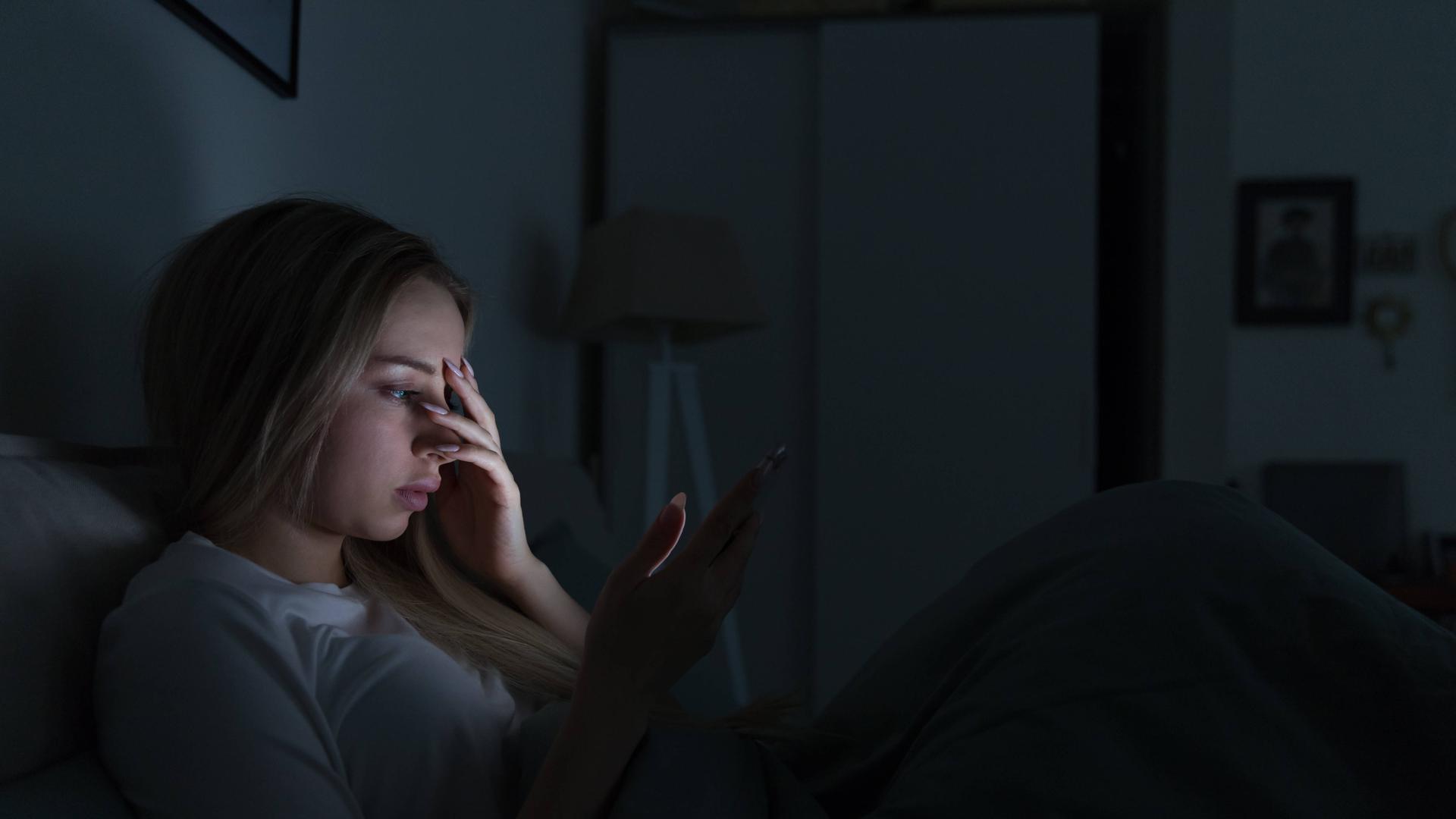 Eine junge Frau liegt in einem dunklen Zimmer in einem Bett. Das Licht ihres Handydisplays erhellt ihr Gesicht. Sie hält sich eine Hand vor das Gesicht. Eine junge Frau liegt in einem dunklen Zimmer in einem Bett. Das Licht ihres Handydisplays erhellt ihr Gesicht. Sie hält sich eine Hand vor das Gesicht.