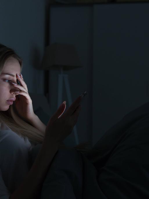 Eine junge Frau liegt in einem dunklen Zimmer in einem Bett. Das Licht ihres Handydisplays erhellt ihr Gesicht. Sie hält sich eine Hand vor das Gesicht. Eine junge Frau liegt in einem dunklen Zimmer in einem Bett. Das Licht ihres Handydisplays erhellt ihr Gesicht. Sie hält sich eine Hand vor das Gesicht.