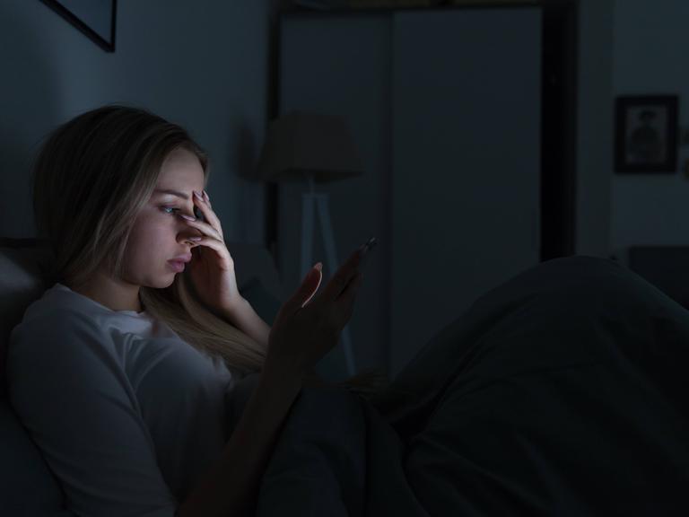 Eine junge Frau liegt in einem dunklen Zimmer in einem Bett. Das Licht ihres Handydisplays erhellt ihr Gesicht. Sie hält sich eine Hand vor das Gesicht. Eine junge Frau liegt in einem dunklen Zimmer in einem Bett. Das Licht ihres Handydisplays erhellt ihr Gesicht. Sie hält sich eine Hand vor das Gesicht.