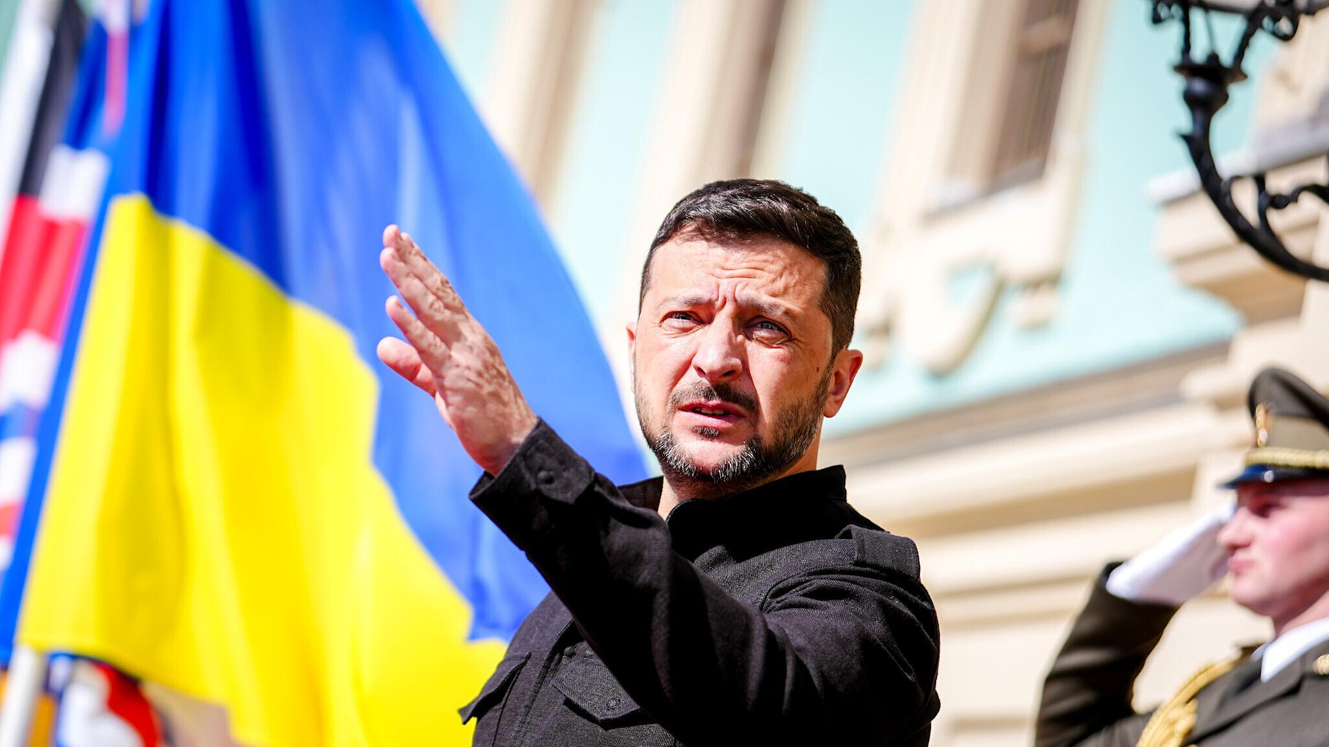 Der ukrainische Präsident Wolodymyr Selenskyj steht mit ausgestreckter Hand vor dem Marienpalast in Kiew; im Hintergrund weht die ukrainische Flagge