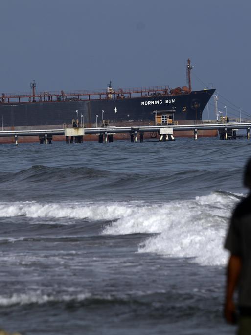 Ein Mann spaziert am Strand von El Palito entlang, im Hintergrund ist die Raffinerie zu sehen, der unter panamaischer Flagge fahrende Öltanker Morning Sun am Dock der Raffinerie El Palito in Puerto Cabello, Bundesstaat Carabobo.
